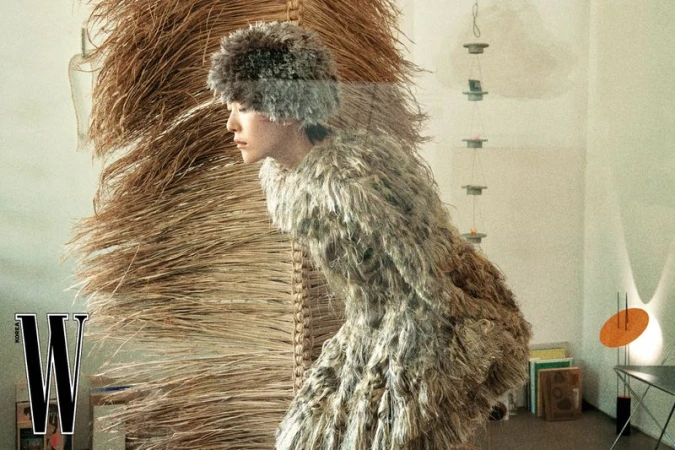 Model wearing a textured fuzzy dress and matching fur hat standing near a large woven straw structure in an antique shop.
