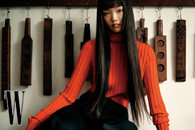 Model with long dark hair wearing a ribbed orange turtleneck against an antique background