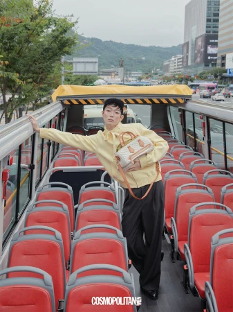 Model in pale yellow jacket holding a structured handbag inside an open top bus with red seats.