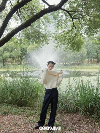 A man wearing a cream knit sweater and dark trousers posing outdoors in a park near a pond with a fountain.