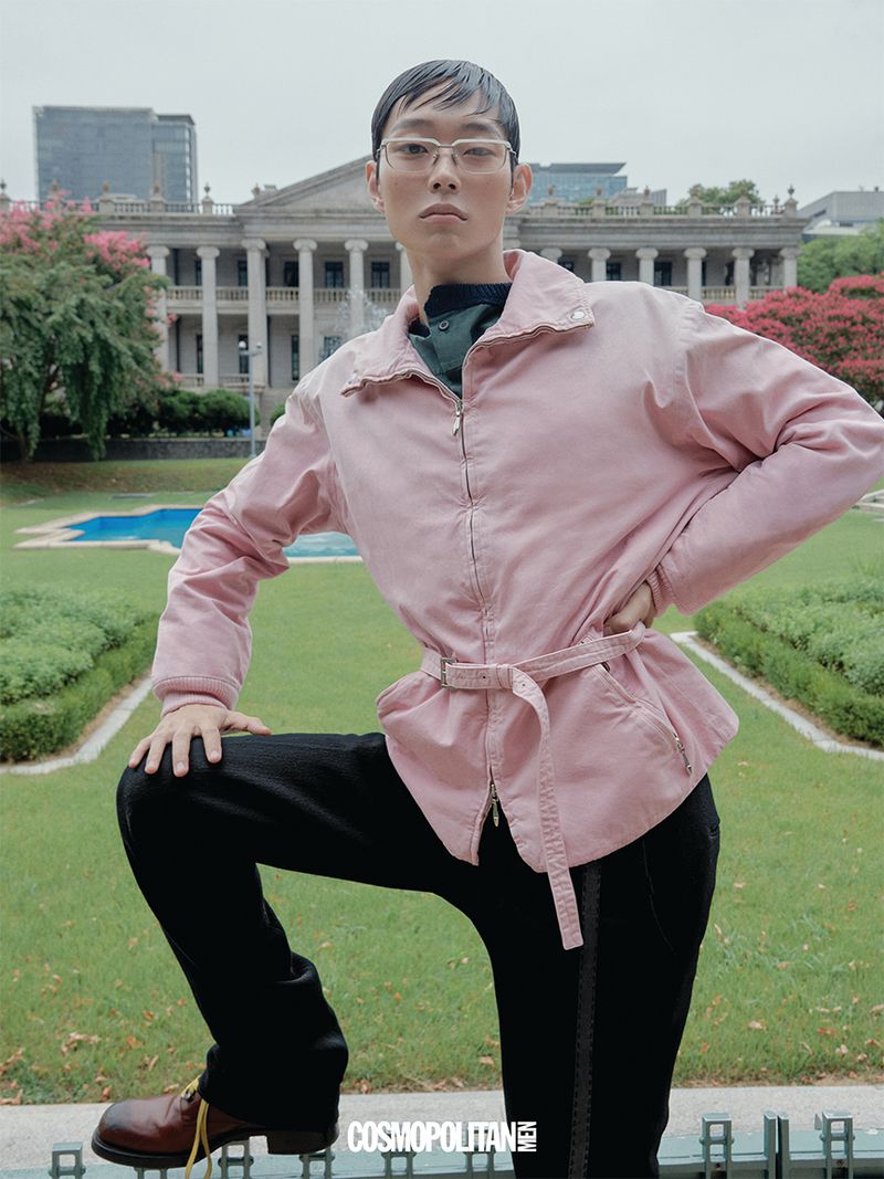Model wearing a pink zippered jacket and black trousers posing in front of a classical stone building with columns.