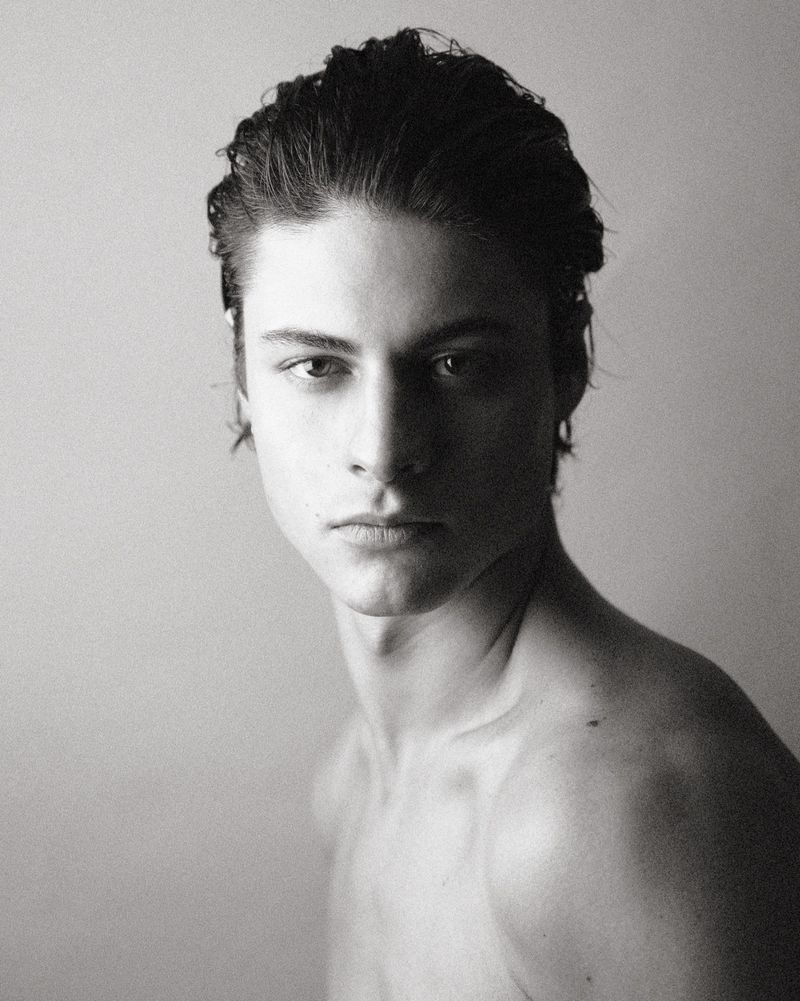 Portrait of a young man with dark wet-styled hair looking directly at the camera against a plain light background.