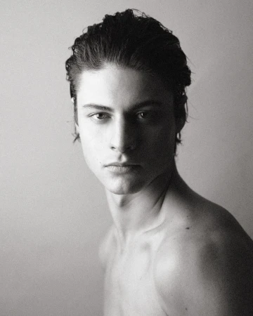 Portrait of a young man with dark wet-styled hair looking directly at the camera against a plain light background.
