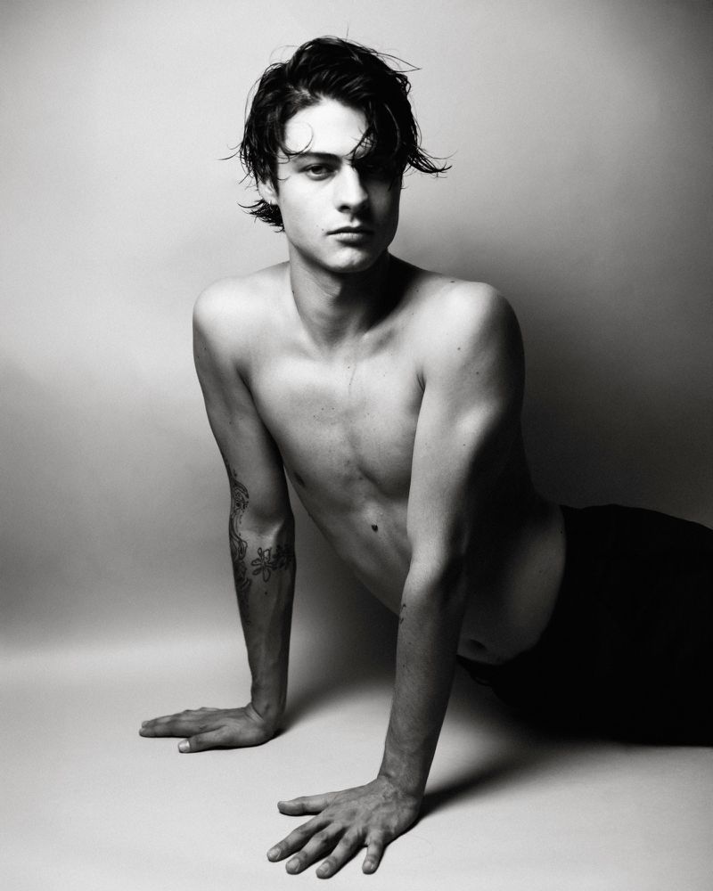 Black and white portrait of a shirtless young man resting on his hands against a seamless studio background