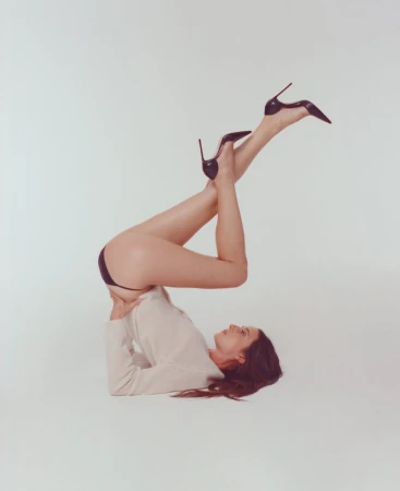 Model lying on back with legs raised wearing a cream top and black stiletto heels against a white backdrop