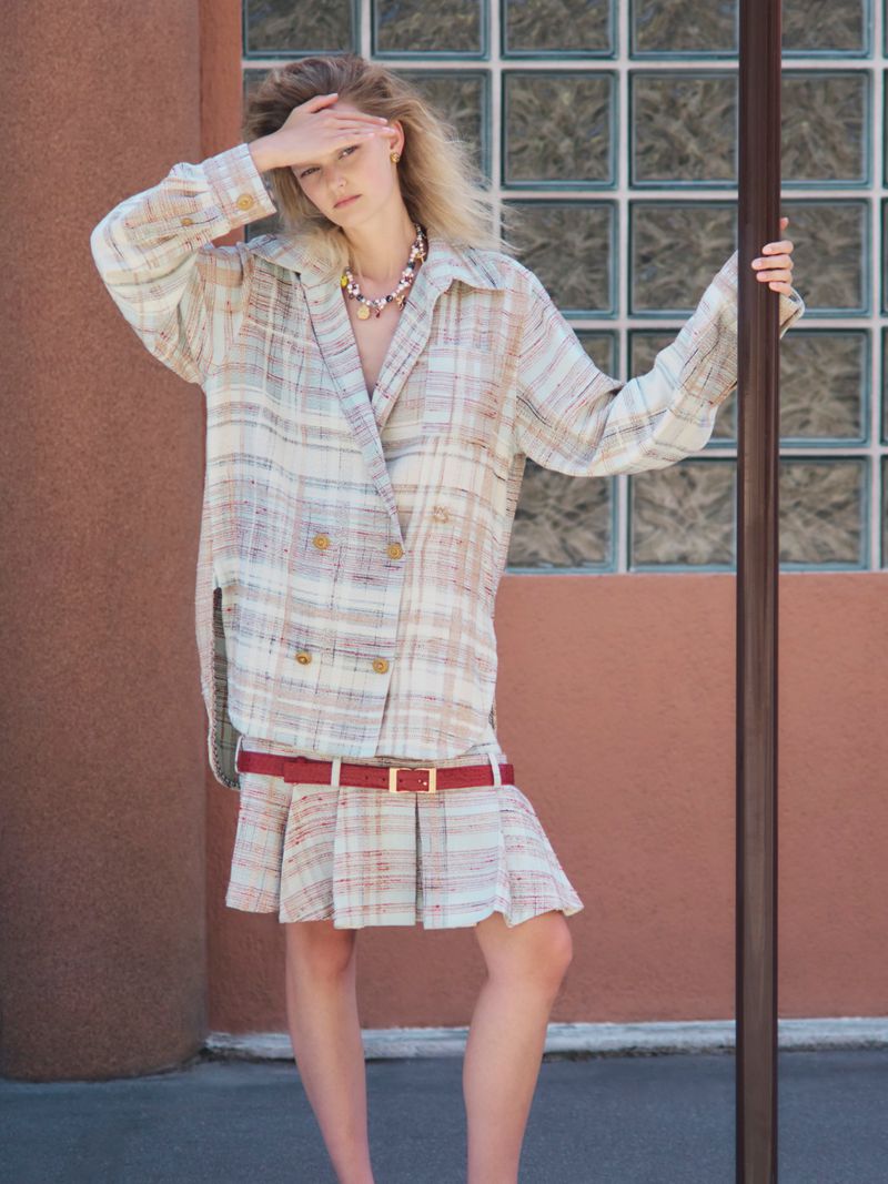 Model in plaid blazer and skirt with red belt holding hand over forehead outdoors against glass block wall.