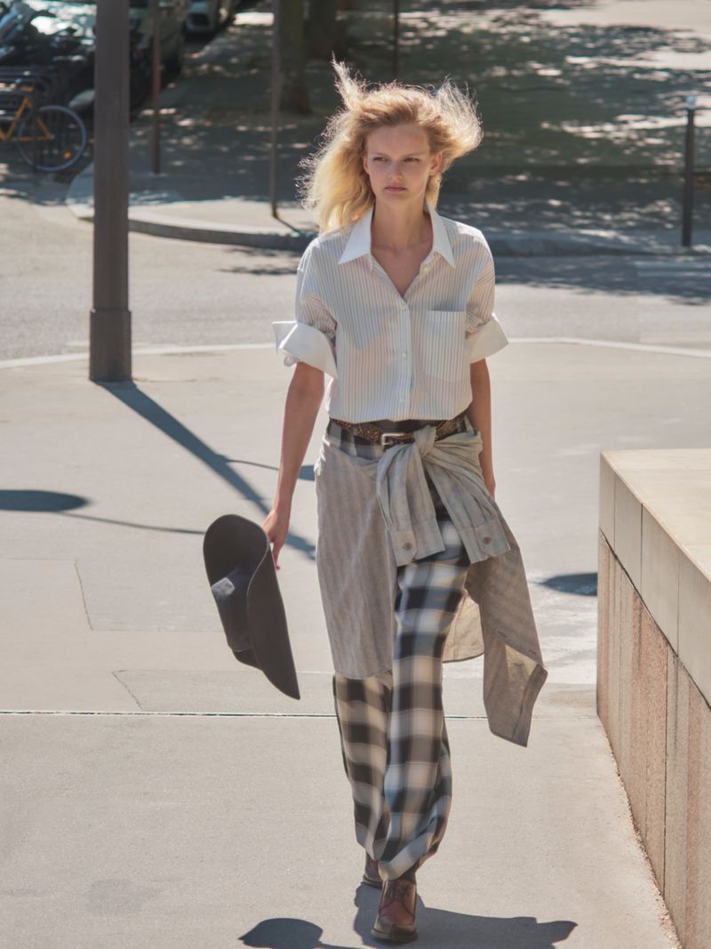 Model walking on an outdoor city street wearing a striped shirt and patterned pants while holding a dark hat.
