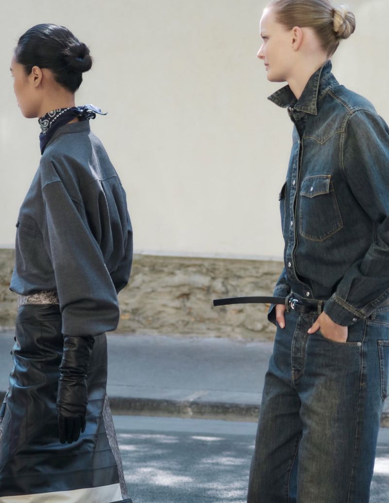 Two female models in denim and dark clothing walking past a pale building exterior.