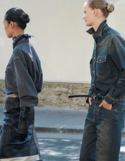 Two female models in denim and dark clothing walking past a pale building exterior.
