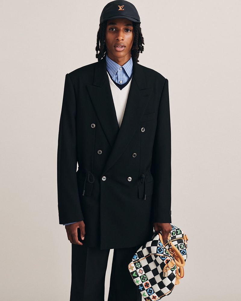 Model wearing a double-breasted black jacket holding a checkered monogram duffel bag against a plain background