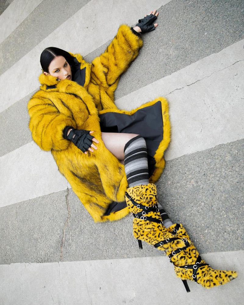 A fashion shot of a young adult woman in a yellow fur coat, striped socks, and leopard-print boots lying on a grey and white striped background.