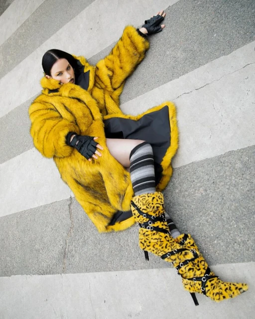 A fashion shot of a young adult woman in a yellow fur coat, striped socks, and leopard-print boots lying on a grey and white striped background.