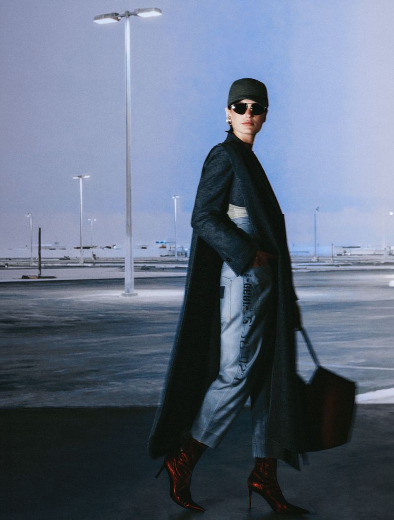 Model in dark coat and cap holding a handbag outdoors under parking lot lights at dusk.