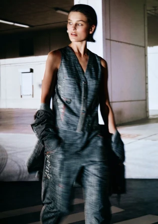 Model wearing a dark grey textured sleeveless vest and trousers walking through a concrete parking structure.
