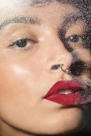 Close-up beauty portrait of a woman with glossy red lips and septum ring viewed through a wet glass pane