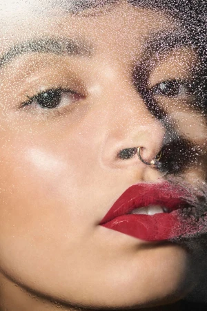 Close-up beauty portrait of a woman with glossy red lips and septum ring viewed through a wet glass pane