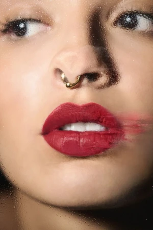 Close-up portrait of a woman wearing red lipstick and a gold septum ring, viewed through a water-streaked glass surface.