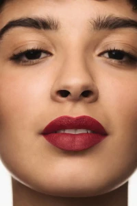 Close-up portrait of a woman wearing rich red lipstick with dark, defined eyebrows against a neutral background.