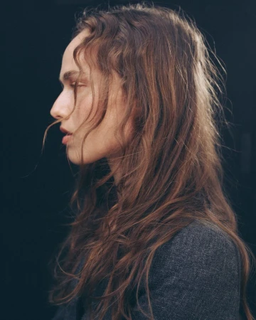 Profile portrait of a woman with long wavy brown hair wearing a dark textured jacket against a plain black background.