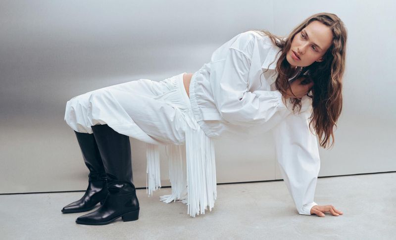 Model in white shirt and fringed pants with black boots posing against a plain grey studio backdrop.