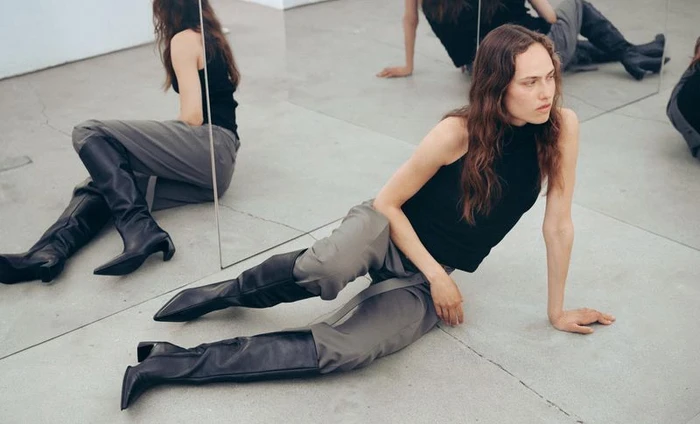 Model seated on concrete floor wearing black sleeveless top, grey trousers and black leather boots against mirrors