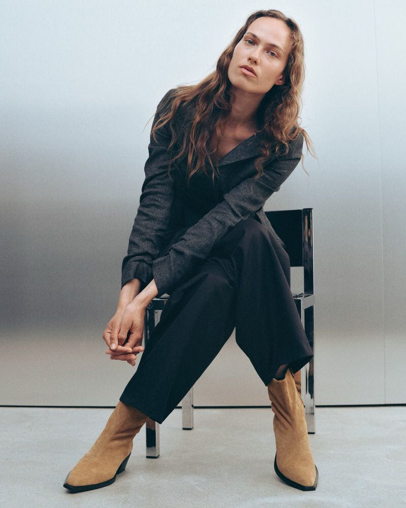 Model Hanna Felding seated on a metallic chair wearing a dark grey jacket, dark trousers, and tan pointed boots.