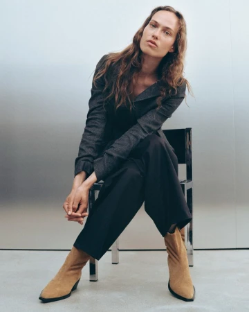 Model Hanna Felding seated on a metallic chair wearing a dark grey jacket, dark trousers, and tan pointed boots.
