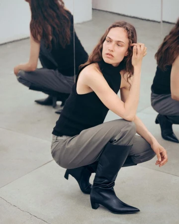 Model Hanna Felding crouching in a black sleeveless top, grey trousers, and black knee-high boots reflecting in mirrors.