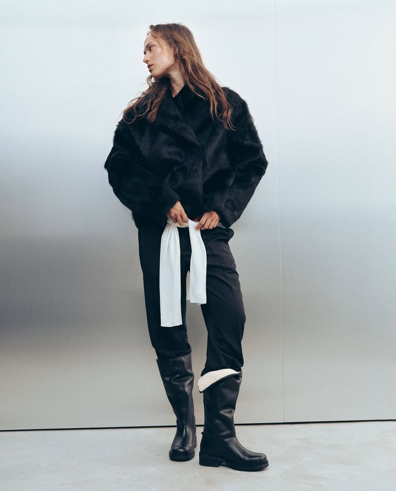 Model standing against a metallic silver background wearing a black textured jacket, black trousers, and tall black leather boots.