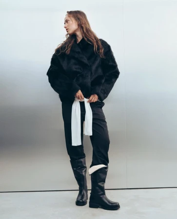 Model standing against a metallic silver background wearing a black textured jacket, black trousers, and tall black leather boots.