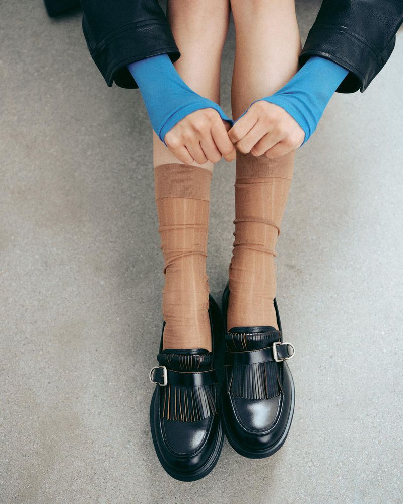 Close-up high-angle shot of a person wearing black leather loafers, sheer tan socks, blue sleeves, and a black jacket against a grey floor