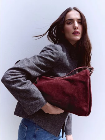 Model Blanca Padilla wearing a grey blazer and holding a burgundy suede shoulder bag against a plain white studio wall