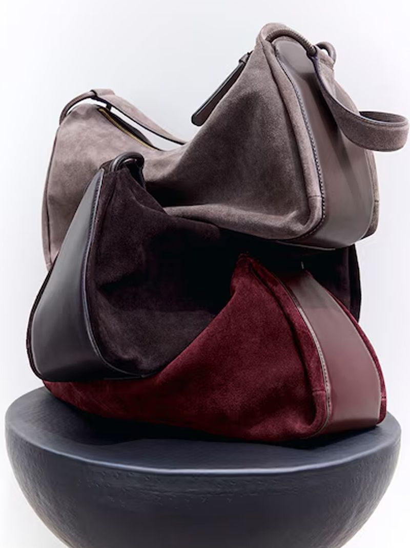 Three stacked suede handbags in brown and burgundy resting on a dark rounded pedestal against a white background