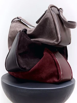 Three stacked suede handbags in brown and burgundy resting on a dark rounded pedestal against a white background