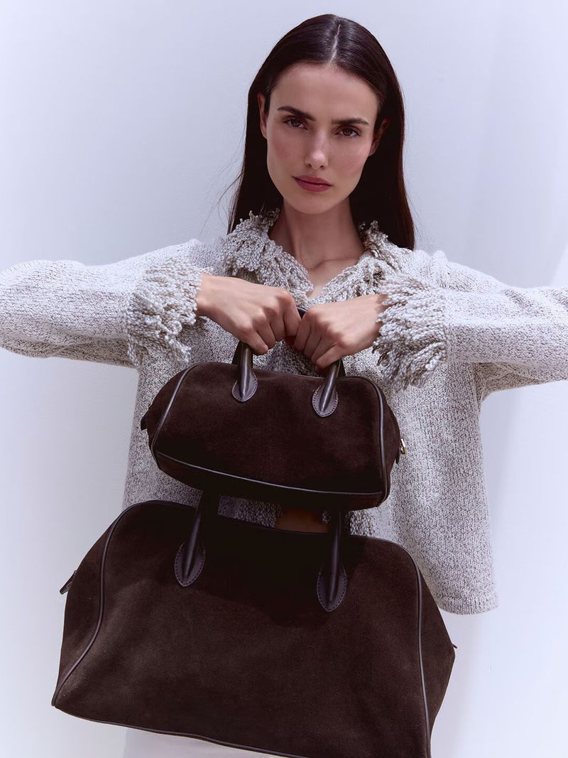 Model Blanca Padilla wearing a textured tweed jacket holding two dark brown leather handbags against a seamless white background