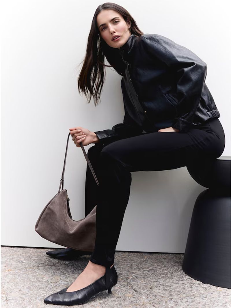 Model Blanca Padilla in a black leather jacket sitting and holding a grey handbag against a white background