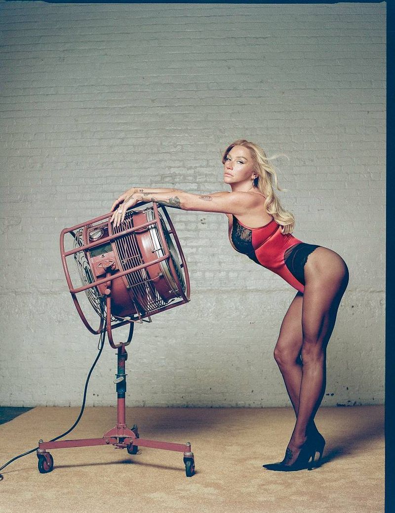 Kesha leaning against a large industrial floor fan wearing a red and black bodysuit and high heels in a studio.