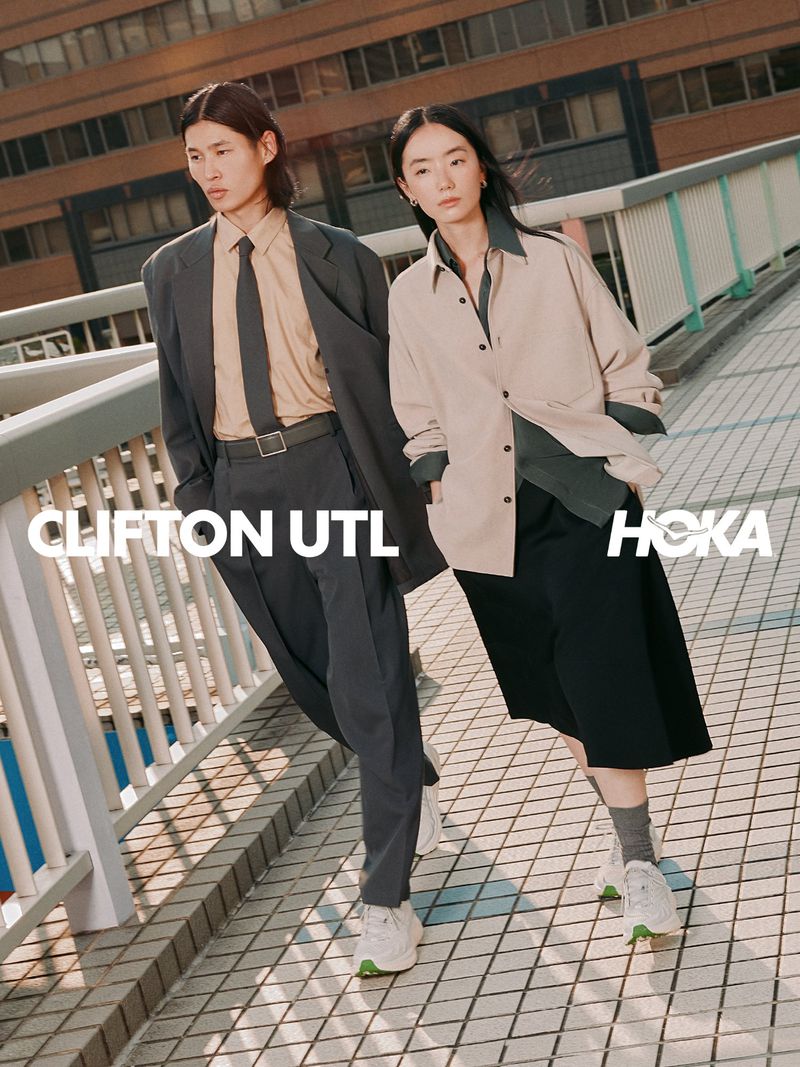 Two models wearing outerwear and sneakers walking outdoors on a tiled bridge walkway with buildings in the background.
