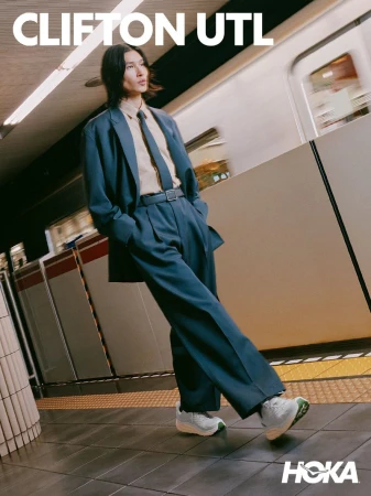 A man wearing a blue suit, tie, and sneakers stands on a subway platform as a train passes blurred in the background.