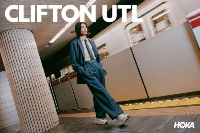 A man in a loose teal suit and sneakers walks on a subway platform next to a train, captured for the HOKA Clifton UTL campaign.