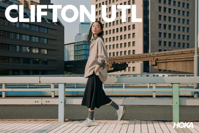 East Asian woman walking outdoors in urban setting wearing beige shirt, black midi skirt, and white sneakers.