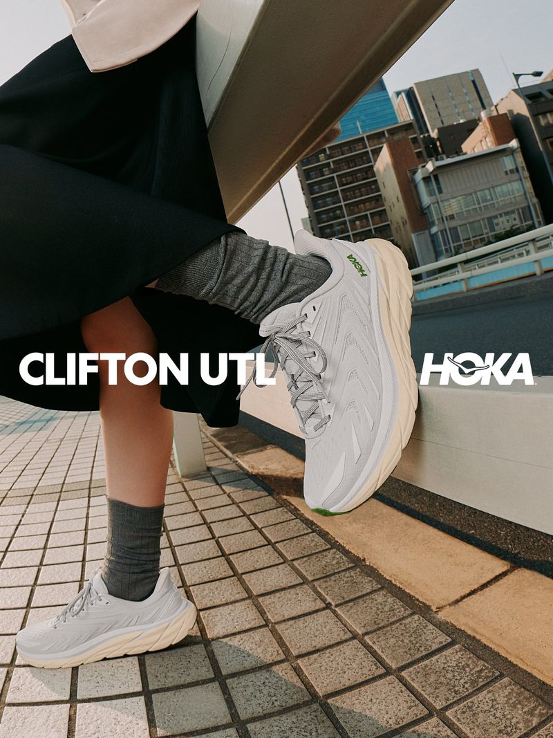 Close-up of a person wearing HOKA Clifton UTL sneakers, grey socks, and a black skirt, resting a foot on an outdoor rail.