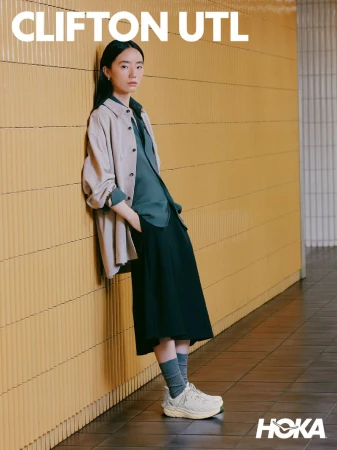 A woman leaning against a yellow tiled wall wearing beige jacket dark shirt black skirt grey socks and white sneakers