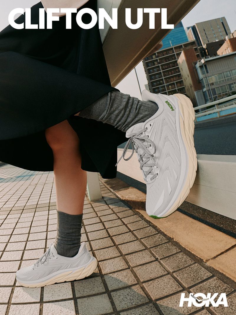 Low angle shot of a person wearing white HOKA sneakers and grey socks outdoors on a tiled rooftop with city buildings in the background.