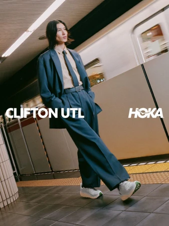 A man wearing a teal suit and white sneakers stands on a subway platform as a train passes by.