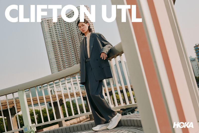 Model wearing an oversized grey blazer and white sneakers leaning against a bridge railing outdoors with tall buildings in the background.