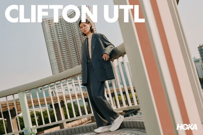 Model wearing an oversized grey blazer and white sneakers leaning against a bridge railing outdoors with tall buildings in the background.