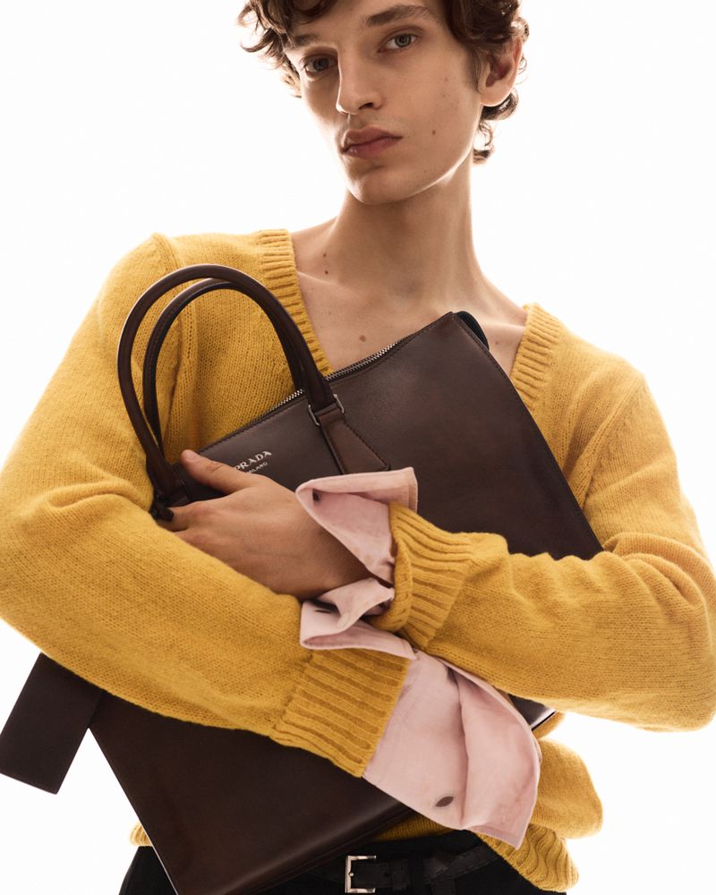 Model in a yellow knit sweater holding a dark brown leather handbag against a bright white background.