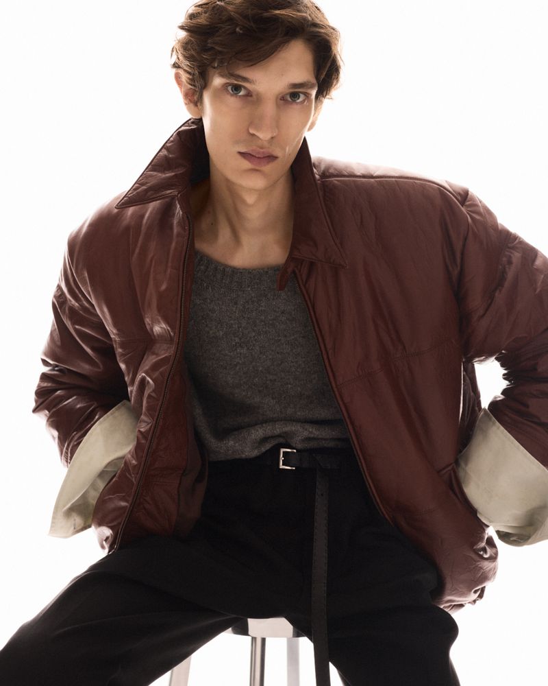 Model wearing a brown leather jacket and dark sweater seated against a bright white background
