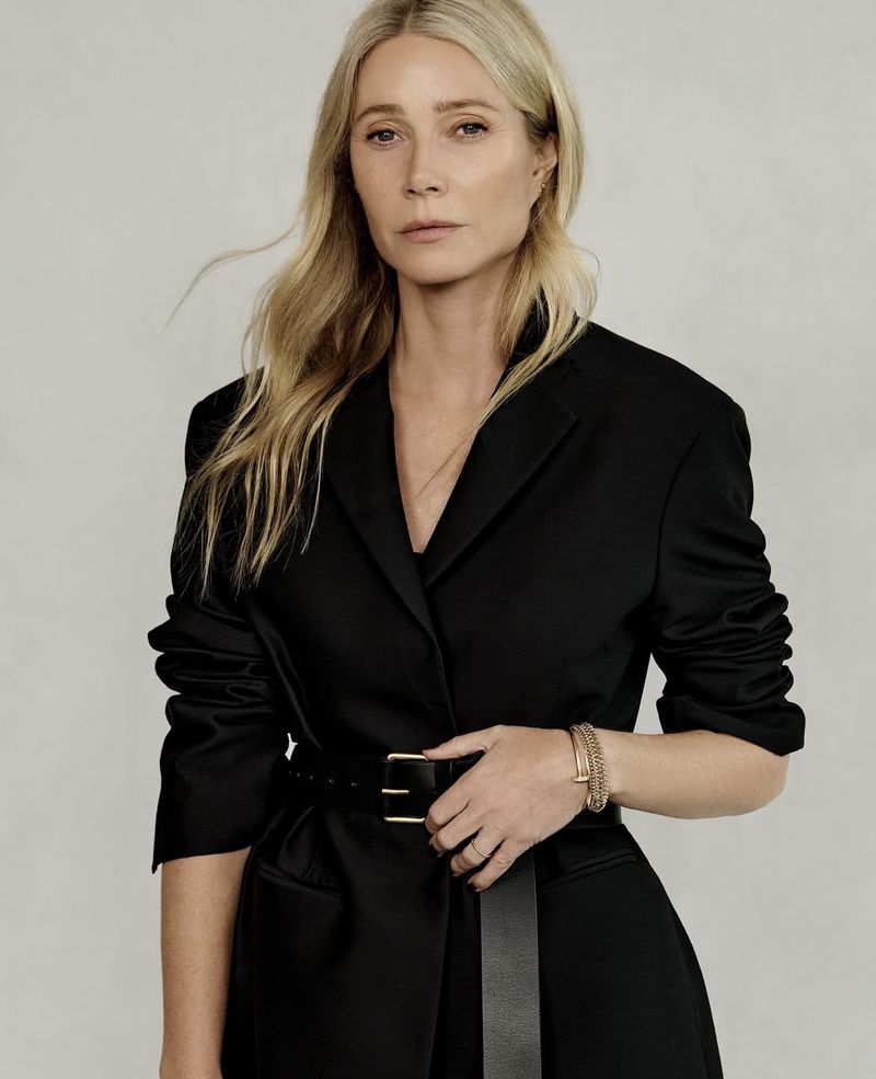 Portrait of a blonde woman in a black blazer cinched with a black belt, wearing bracelets and a ring, posing indoors against a beige background.
